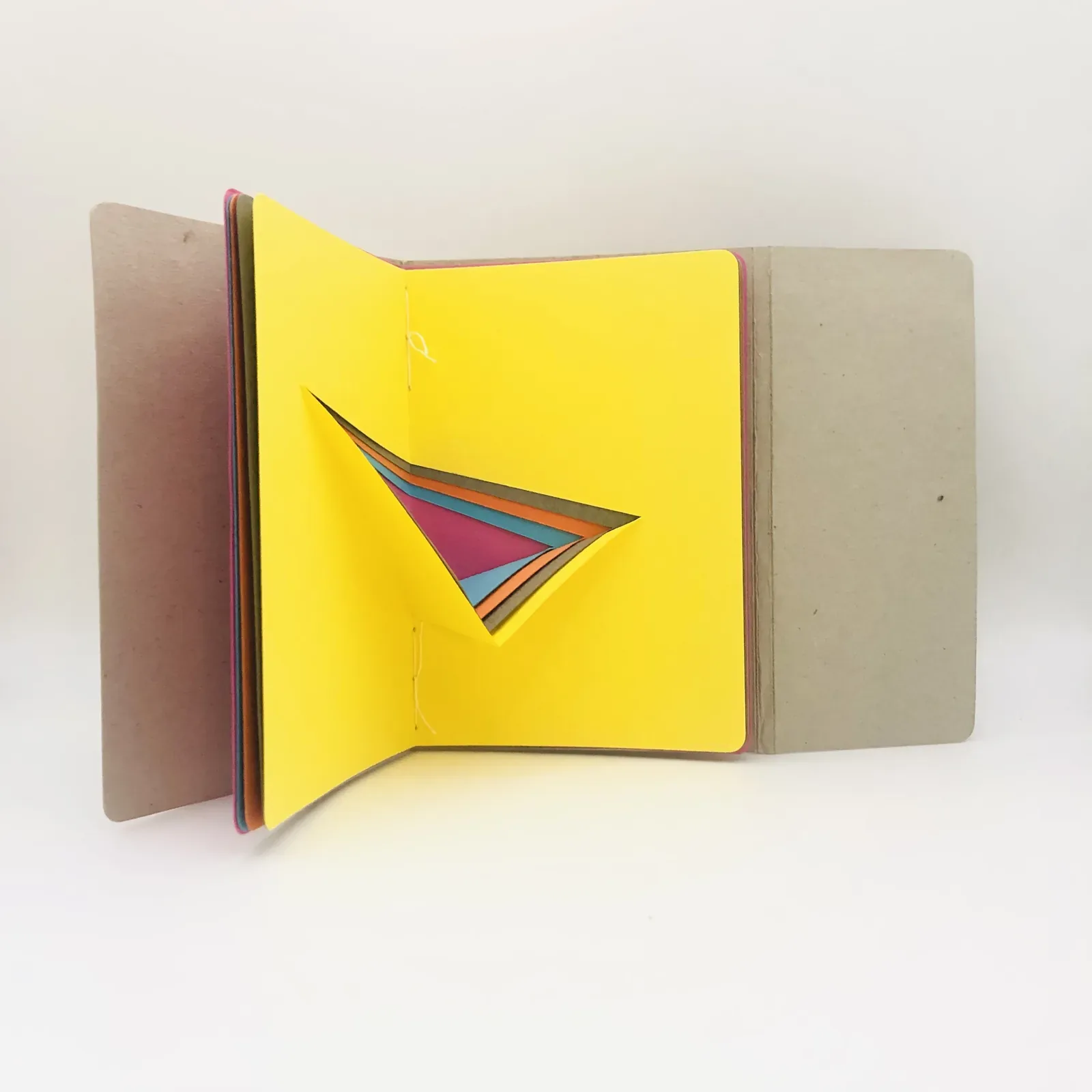 Le carnet ouvert et posé debout : les plats de carton gris s’écartent, la page jaune s’ouvre et la pointe pliée se déploie en rose, orange et turquoise