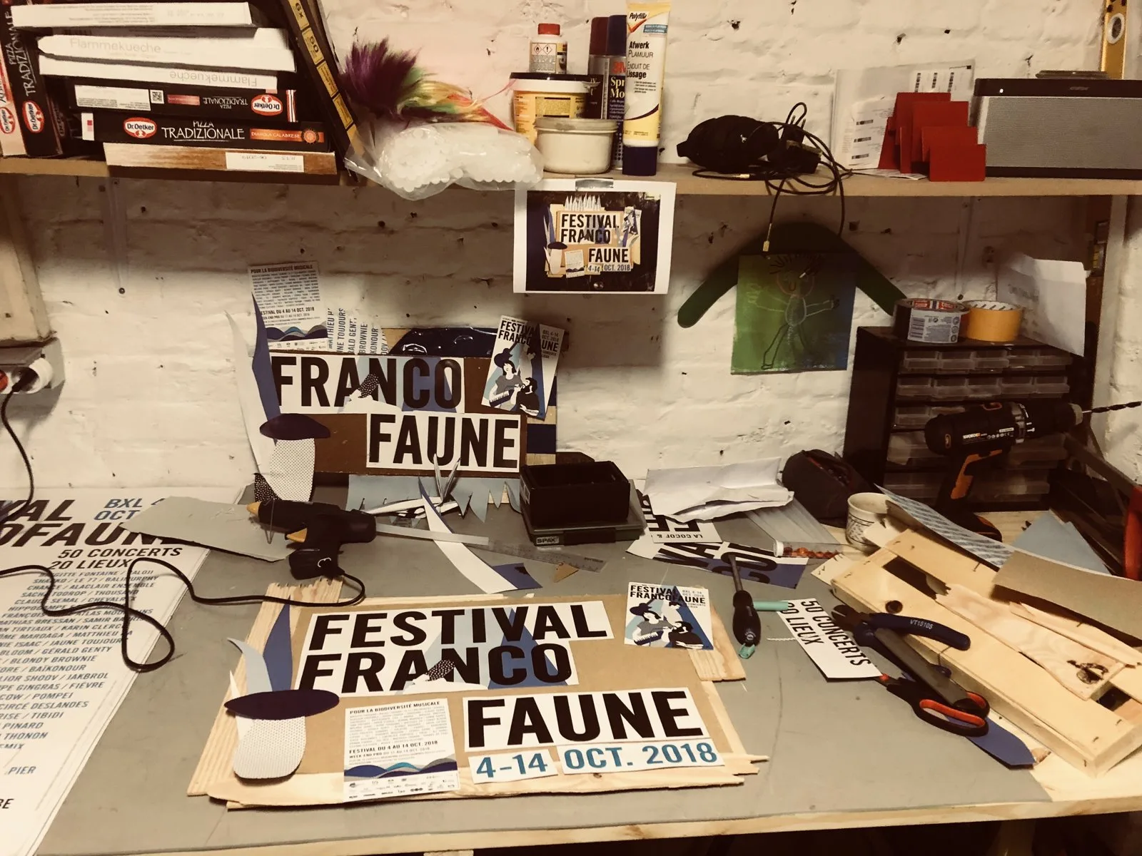Établi encombré d’outils, de pinces et de bocaux, avec des affiches du festival en cours de découpe.