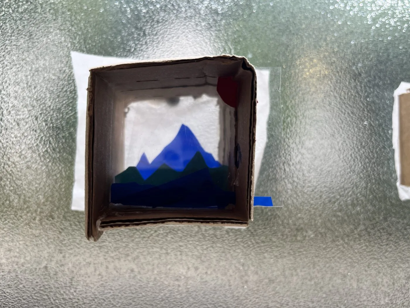 Un cadre de carton posé sur une vitre dépolie : une montagne bleue en papiers découpés