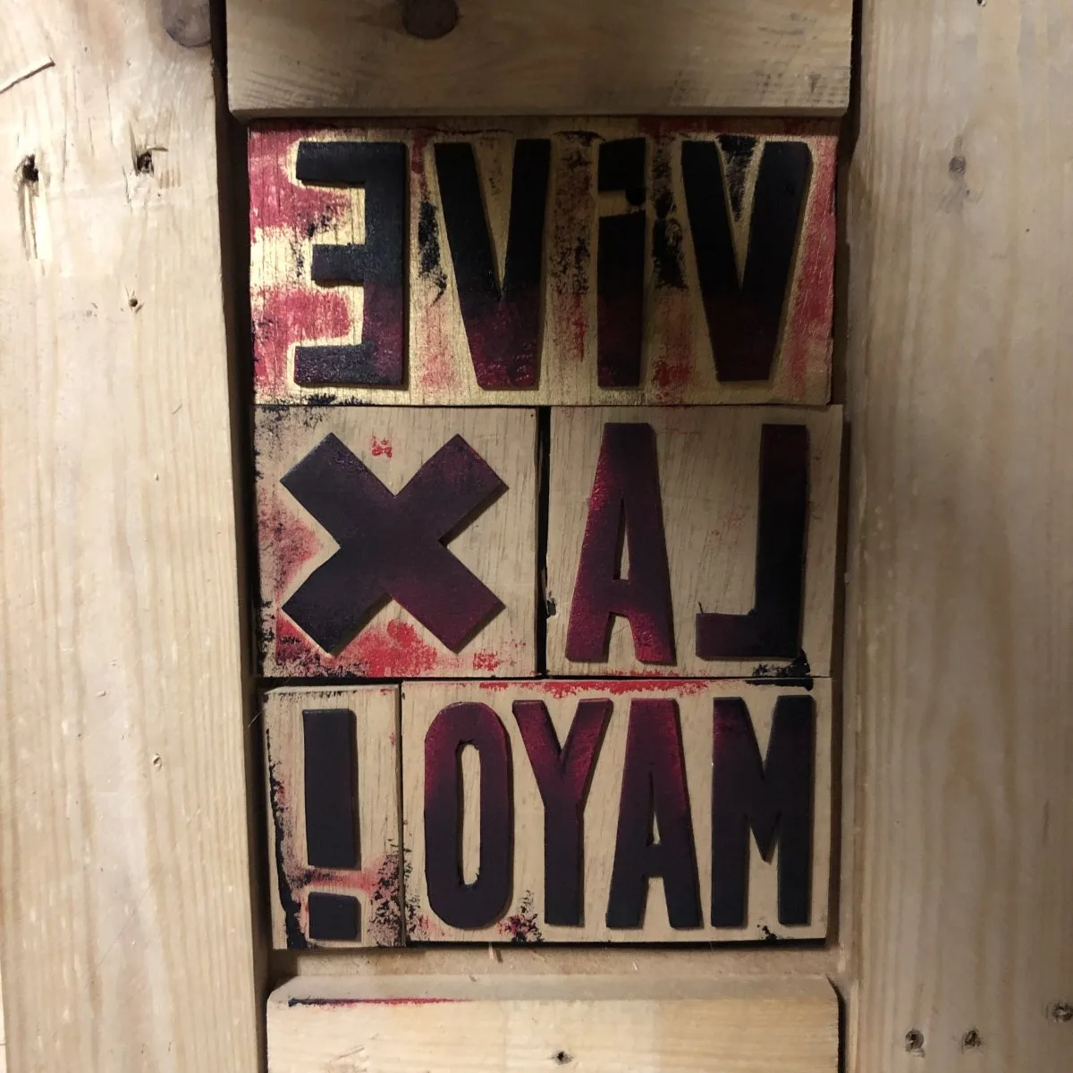Tuiles de lettres encrées de noir et de rouge, assemblées en miroir dans un cadre de bois : VIVE LA X MAYO !