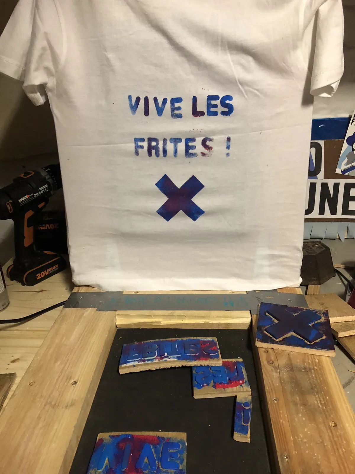 Un t-shirt tiré à la presse — VIVE LES FRITES ! et une croix bleue — encore posé sur la table de tirage