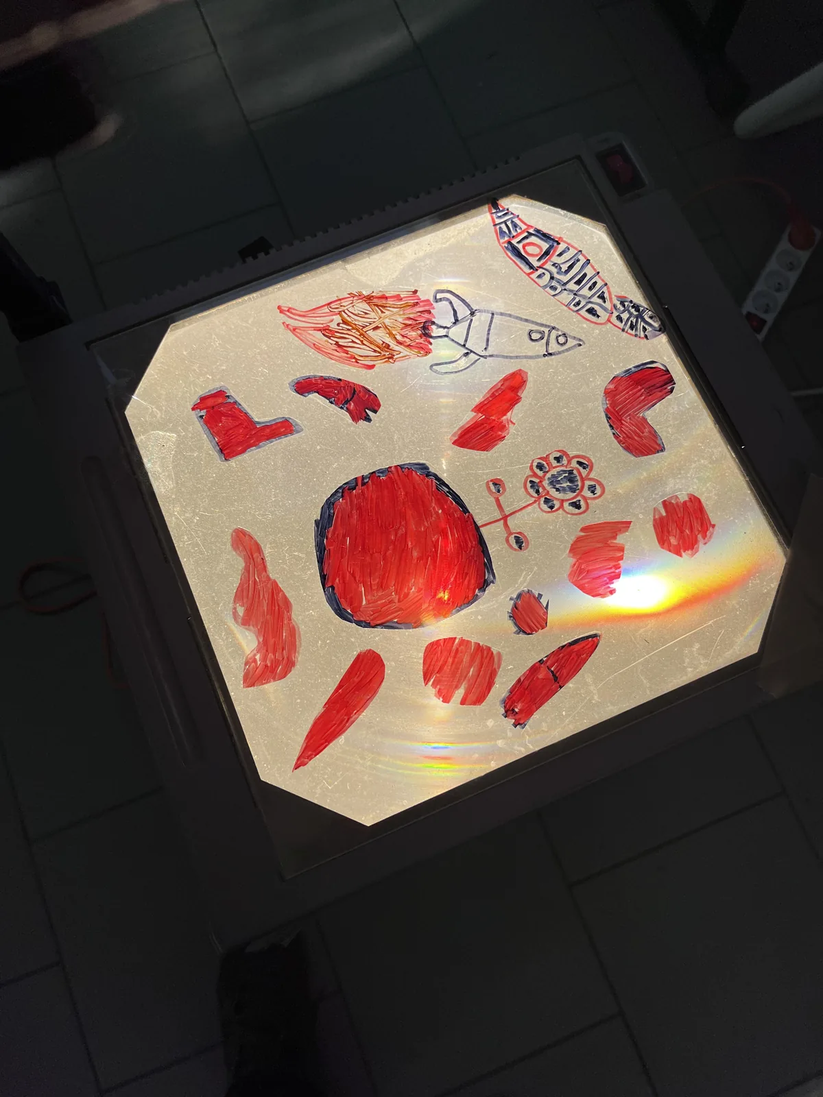Une projection en couleurs — formes rouges et roses translucides posées sur la vitre du rétroprojecteur